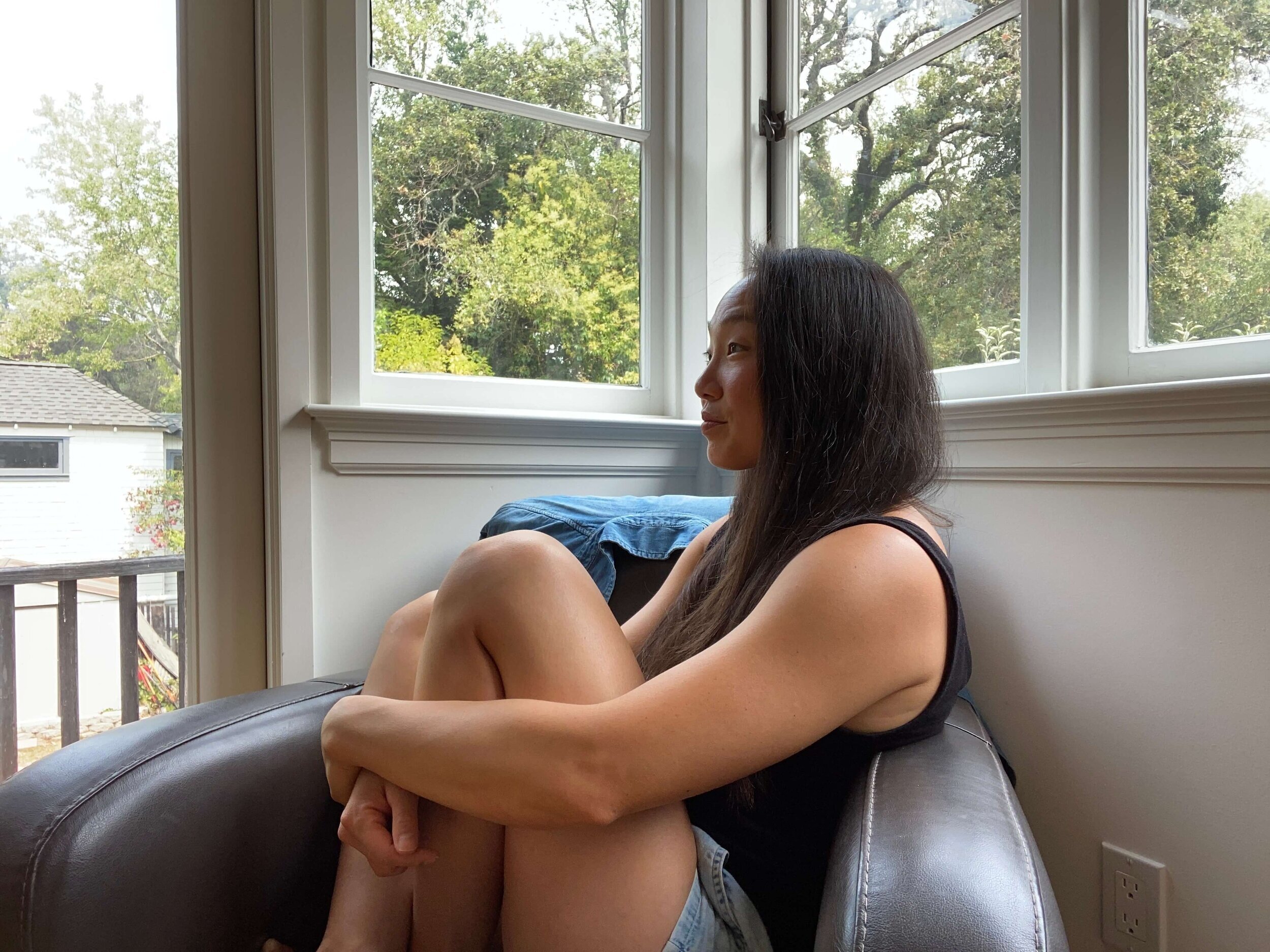 Nina Jung sitting by a window, knees drawn up, looking out at the trees beyond.